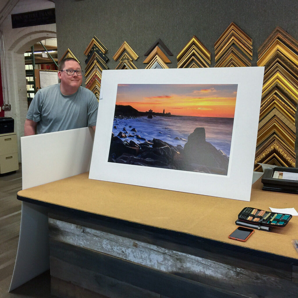 Mike Dooley standing beside a large matted fine art print at Providence Picture Frame, smiling proudly.