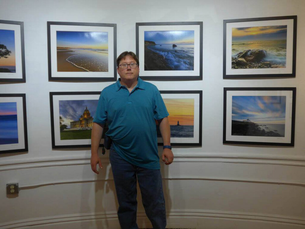 Mike Dooley standing in front of some of his photos at a fine art gallery exhibit of his landscape and seascape prints.