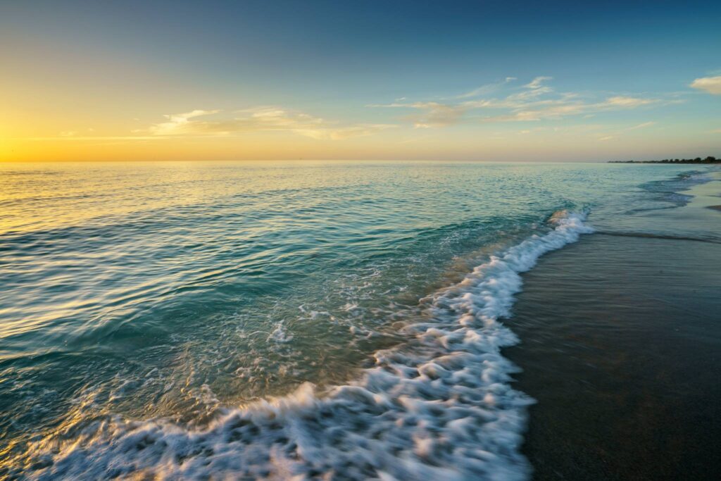 Gentle ocean wave rolls onto a tranquil beach at sunrise, with golden light reflecting on the water and soft clouds above the horizon.