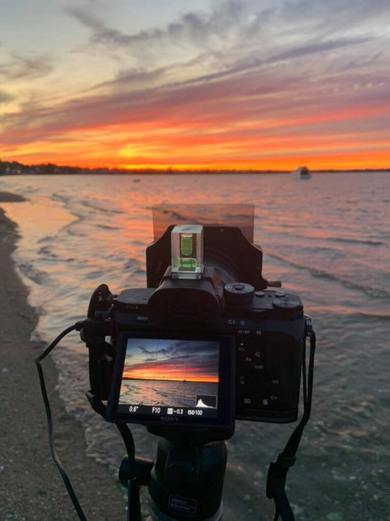 Camera set up at Conimicut Point in Warwick, equipped with a spirit level, filters, and remote release, aimed at the boat “Stugots” anchored against a glowing sunset over the water.