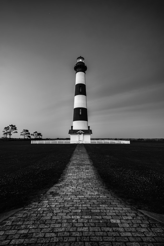 Black and white photograph of Bodie Island Lighthouse standing tall against a dramatic sky, with textured sand and coastal grasses in the foreground.