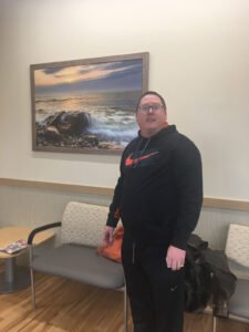 Photographer Mike Dooley standing with a large framed landscape print in a doctors waiting room in East Providence, Rhode Island.