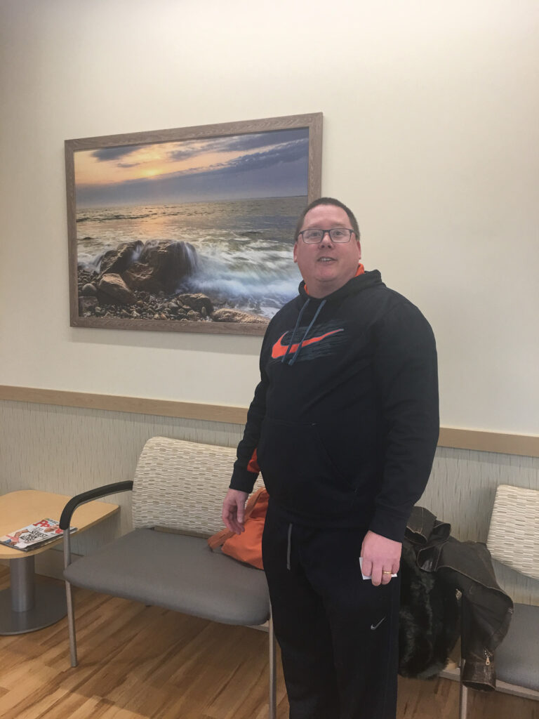 Photographer Mike Dooley standing with a large framed landscape print in a doctors waiting room in East Providence, Rhode Island.