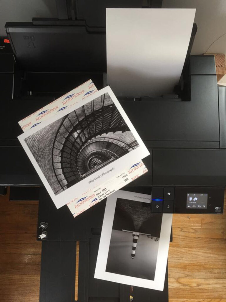Stacks of fine art photography prints by Mike Dooley sitting on the printer, showcasing multiple images ready for framing and display.