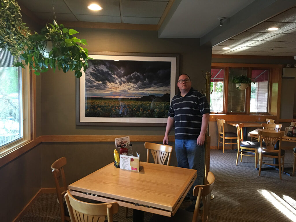 Photographer Mike Dooley standing inside Greggs Restaurant in North Kingstown, holding a large framed print of sunflowers under stormy skies.