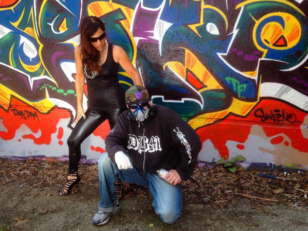 Workshop model and an anonymous graffiti artist standing in front of a colorful graffiti-covered cement wall during a portrait photography session."