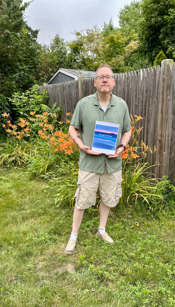 Photographer Mike Dooley standing in his backyard holding a copy of his photography book Through My Eyes.