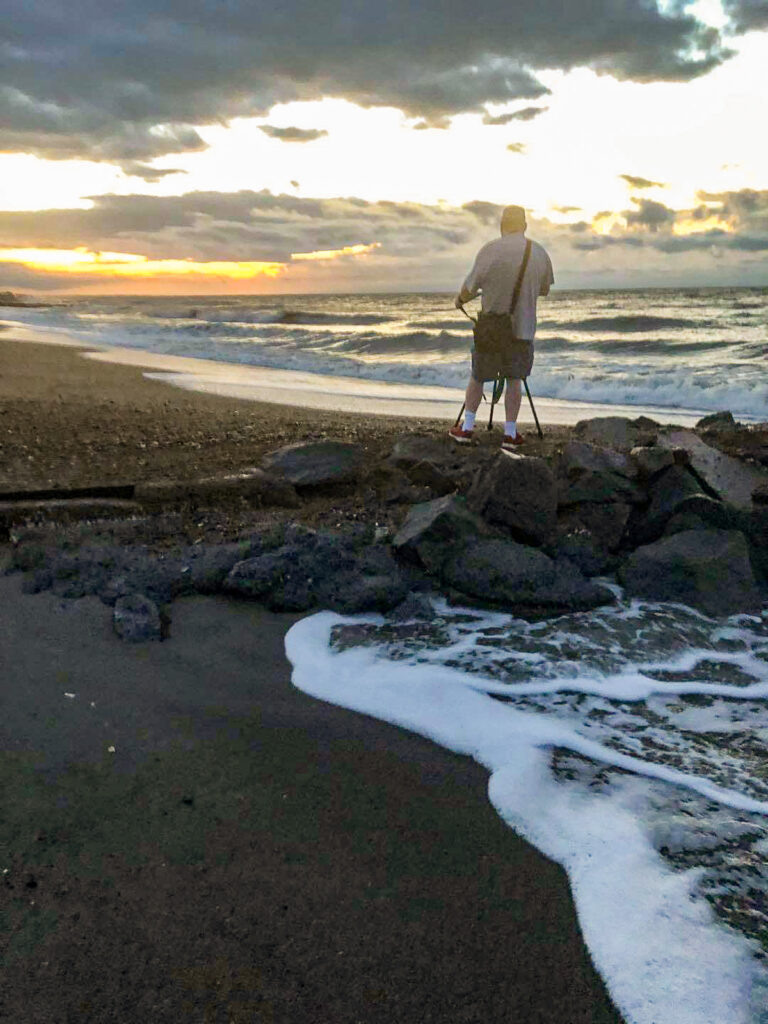 Mike Dooley, seascape photographer, standing on a sandy beach with a camera bag over his shoulder and tripod in front of him, capturing coastal photography.