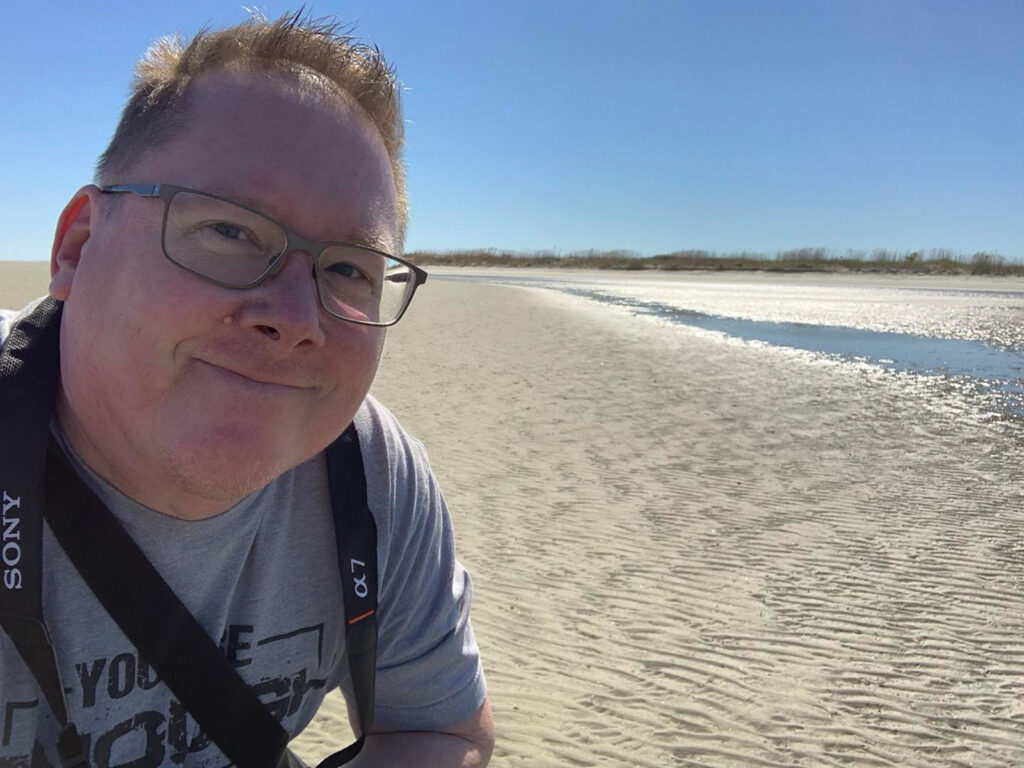 Mike Dooley standing on a sandy beach with the Atlantic Ocean and blue skies in the background, representing his seascape photography work.