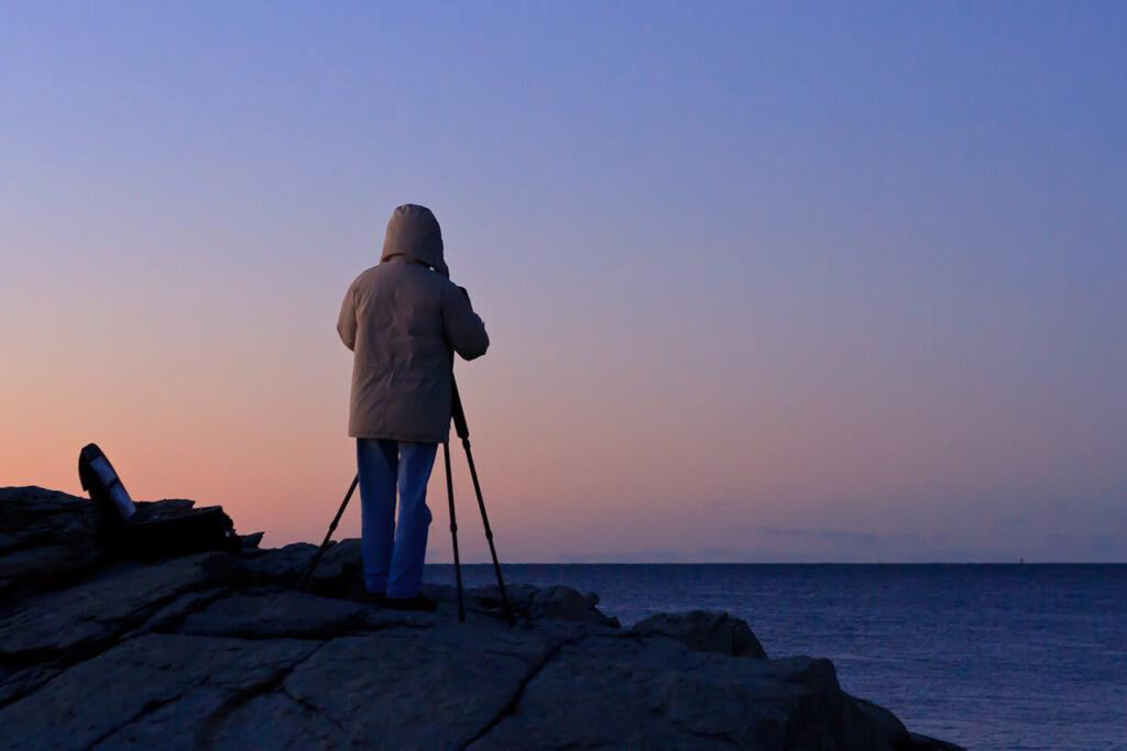 A student standing on rocky cliffs along the Newport coastline, adjusting their tripod and camera to capture the rugged seascape.