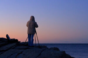 Photography workshop participant standing behind a tripod on the rocky shoreline overlooking Narragansett Bay during a seascape photography class with Mike Dooley Photography.