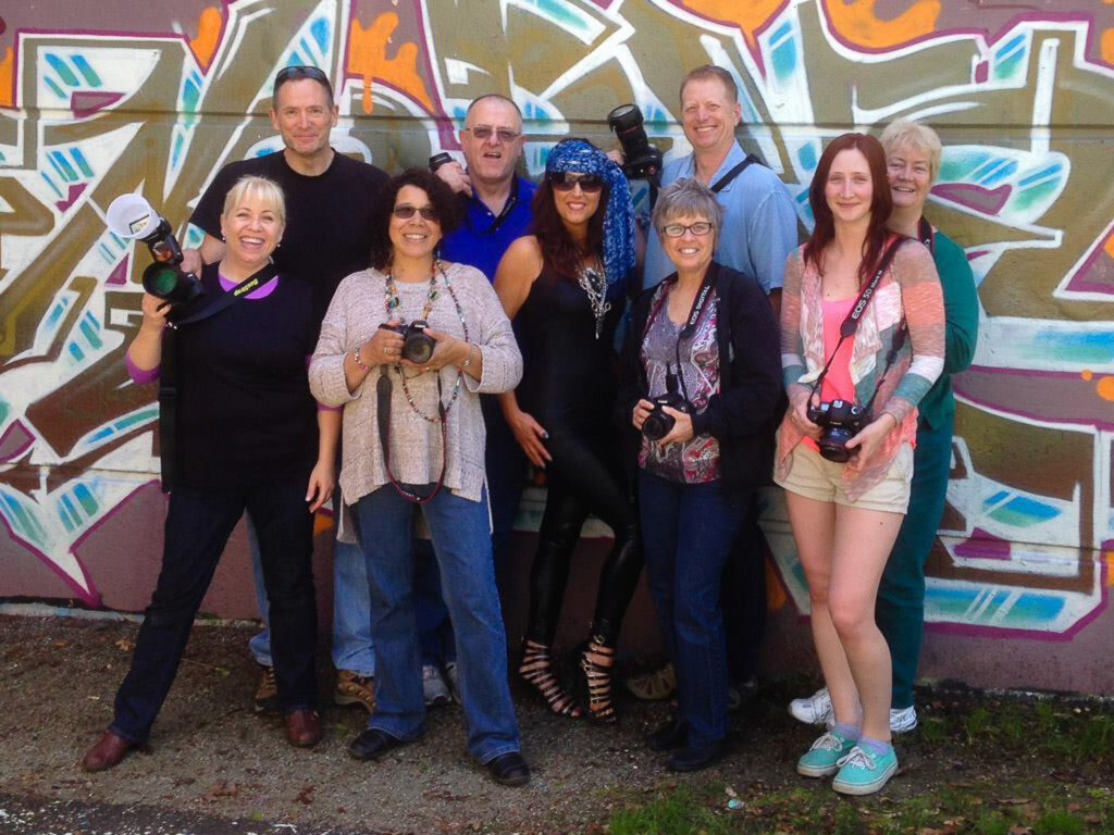 Group of photography students with cameras in hand posing in front of a vibrant graffiti wall during a hands-on lighting workshop.