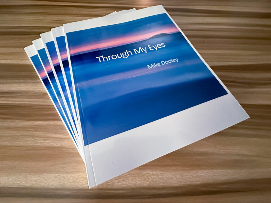 Five copies of 'Through My Eyes' photography book by Mike Dooley, showcasing seascapes and New England landscapes on a table.