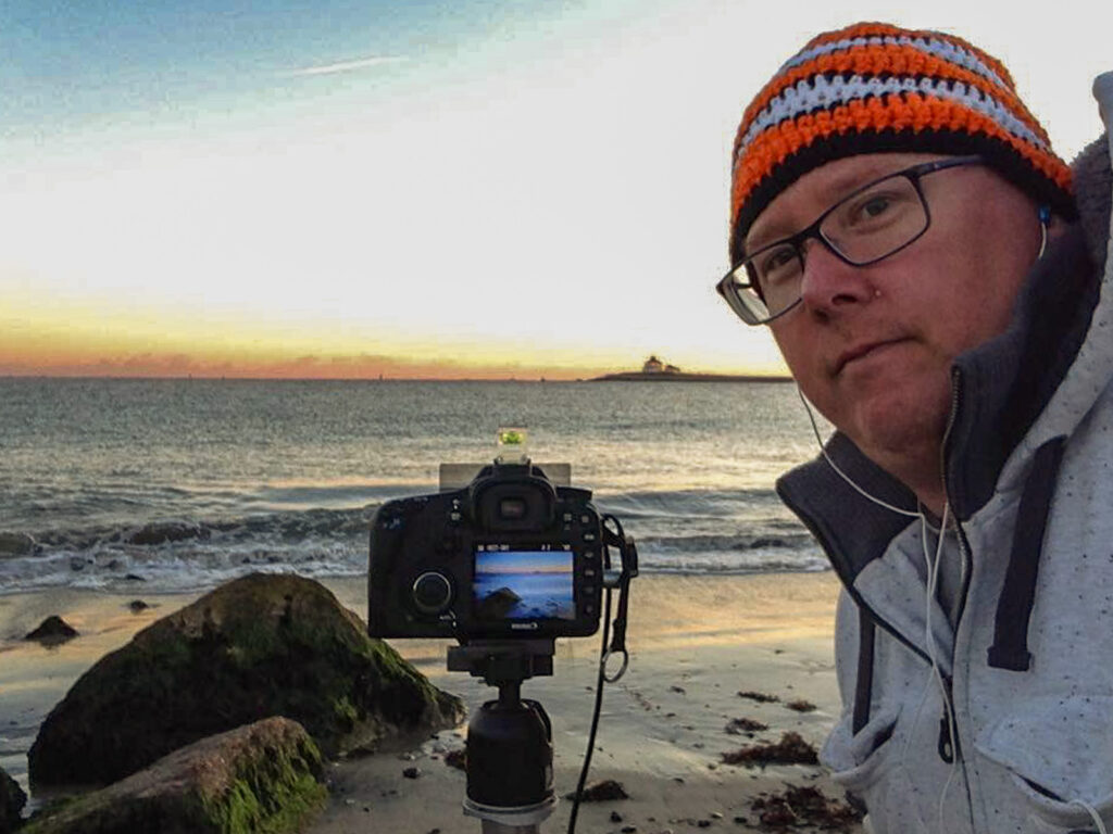 Photographer on a winter beach wearing an orange knit hat with a tripod set up in the sand