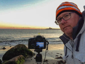 Photographer Mike Dooley on a winter beach wearing an orange knit hat with a tripod set up in the sand