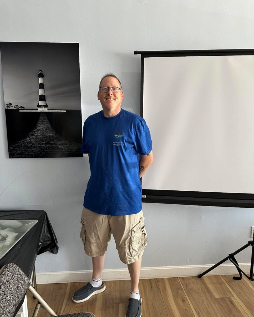 Mike Dooley standing next to a 24x36" metal print of Bodie Island Lighthouse at his Through My Eyes book signing, showcasing the fine art photograph in black and white.