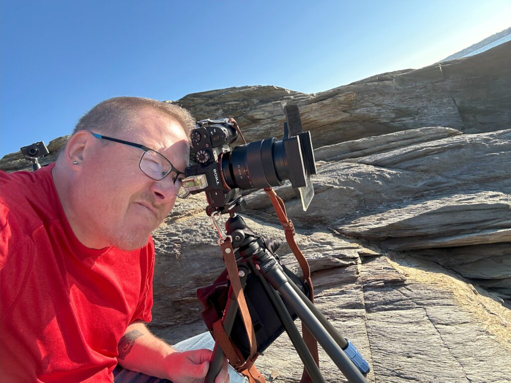 Mike Dooley lying on rocks with tripod, filters, and spirit level, taking a low-angle photograph of the New England coast.