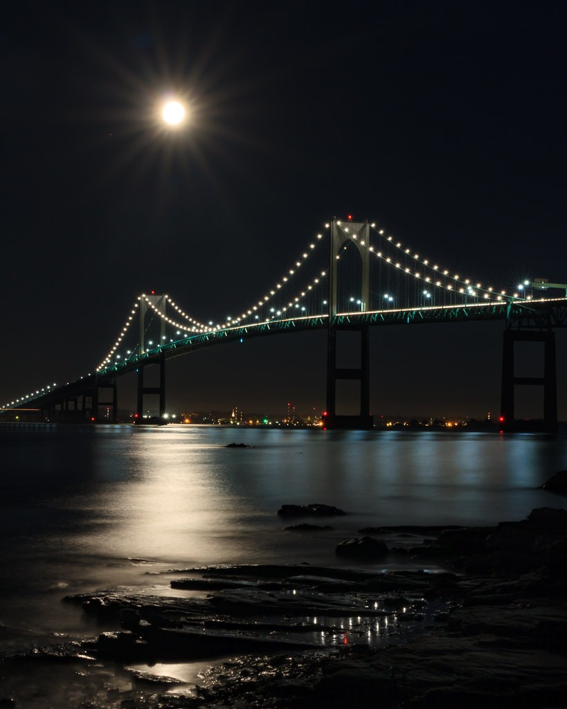 Stunning night photography of a blue moon rising above the Newport Bridge, with reflections shimmering on the water and dramatic low light composition.