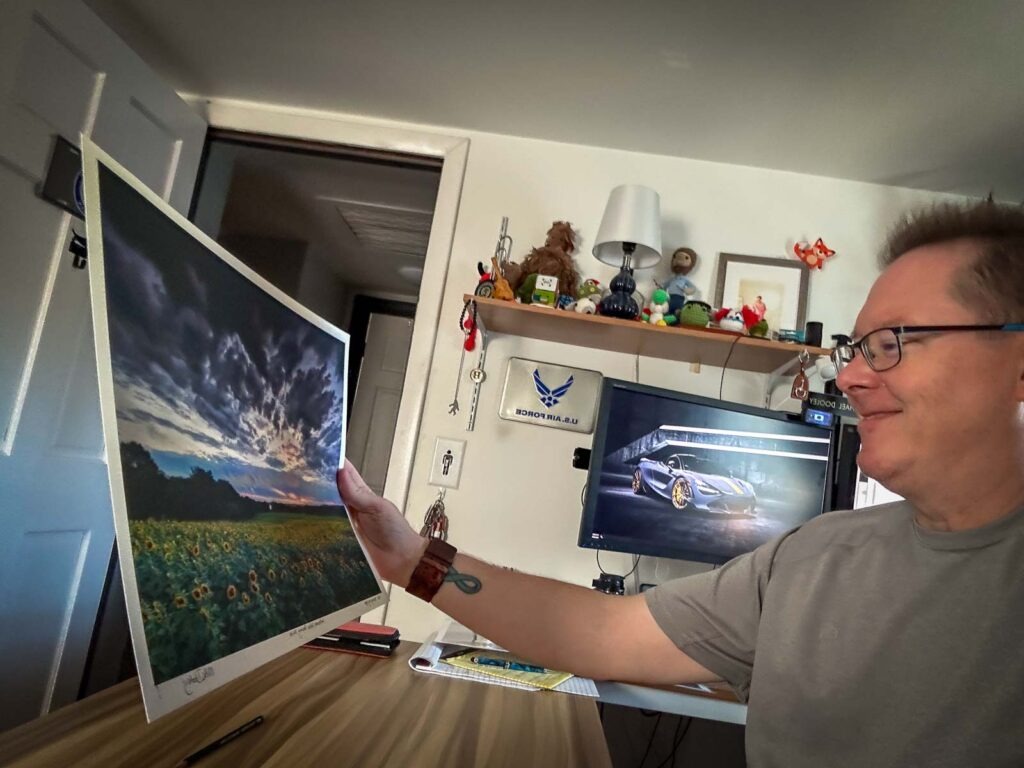 Photograph of Mike holding a signed, numbered 13x19 print titled “Sunflowers Under Stormy Skies,” featuring golden blooms beneath dramatic storm clouds; part of the Signature series.