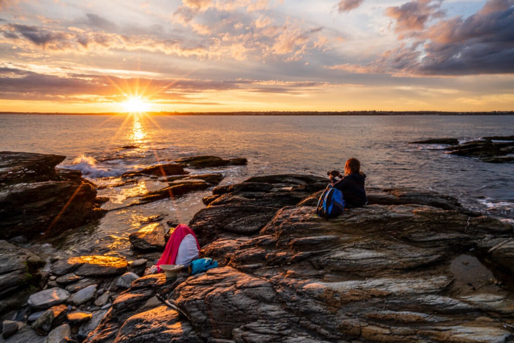 Photography students on the rocky shoreline at Beavertail State Park during sunset over Narragansett Bay, Rhode Island, capturing the warm evening light.