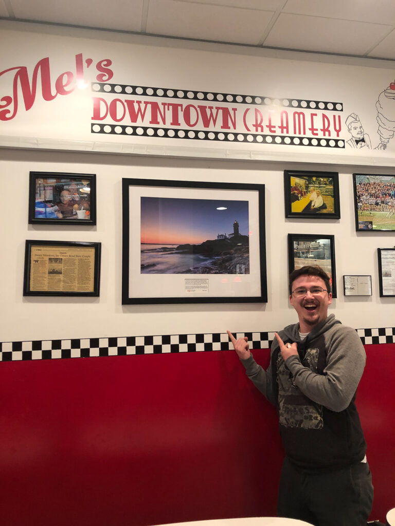 Young man smiling with a Mike Dooley lighthouse print of Beavertail State Park hanging on an ice cream shop wall in Connecticut.