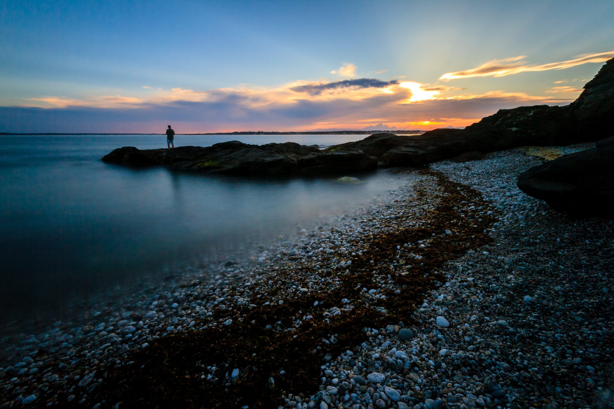 A fisherman standing on rocks at Beavertail State Park in Narragansett Bay, Rhode Island, casting a line, photographed by Mike Dooley.