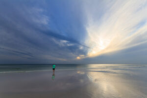 A man walking alone on a beach at sunset, with warm sunlight reflecting on the sand and water, photographed by Mike Dooley.