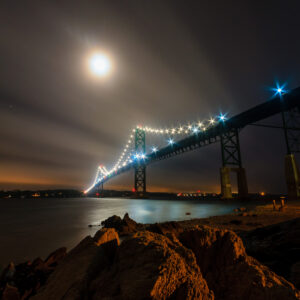 Mount Hope Bridge at night, with clouds moving and moonlight reflecting on the structure and water, photographed by Mike Dooley.