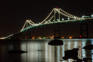 The Newport Bridge illuminated at night over calm waters, with stars in the sky, photographed by Mike Dooley.