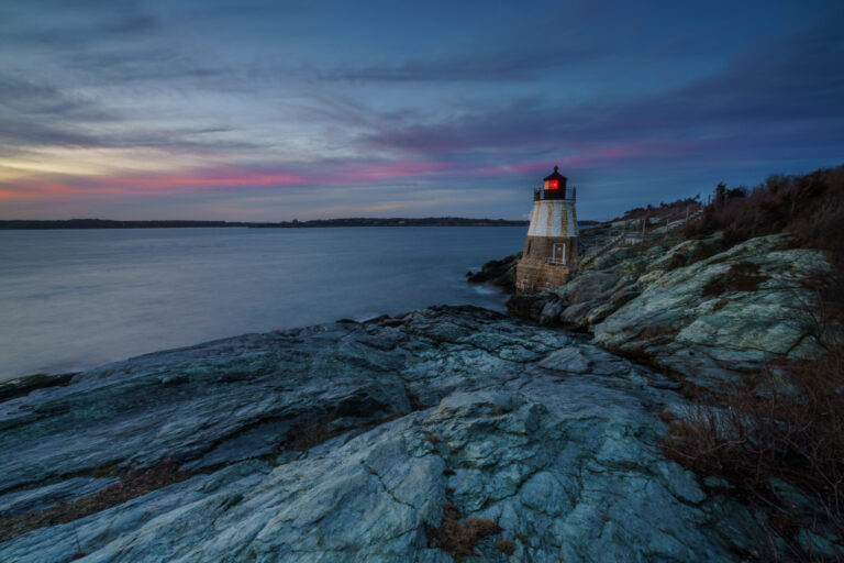 Castle Hill Lighthouse at sunset with its red light on under pink-streaked clouds, rocky foreground, Newport, Rhode Island, by Mike Dooley