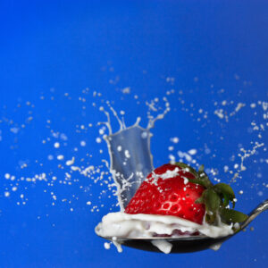 Strawberry falling into a spoonful of milk against a blue background, photographed as a fine art print by Mike Dooley.
