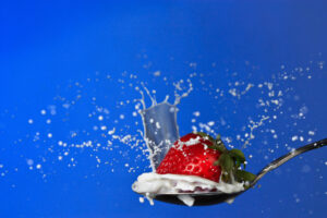 Strawberry falling into a spoonful of milk against a blue background, photographed as a fine art print by Mike Dooley.