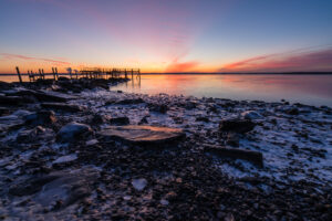 Frozen rocks and icy shoreline at sunrise, with the old Rocky Point pier pilings against a pink and orange sky in Warwick, RI