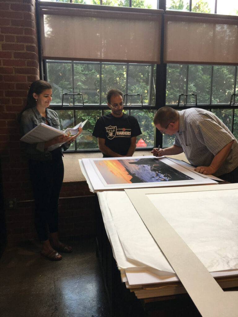 Mike Dooley signing a fine art print on paper, with the art director and print framer looking on, ensuring quality and authenticity.