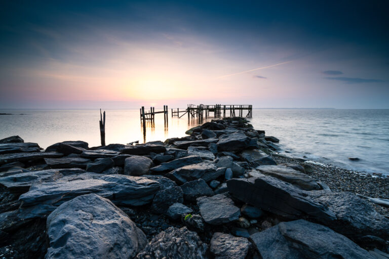 Old pier pilings beyond dark shoreline rocks under a soft blue and pink sky at Rocky Point, Warwick, Rhode Island, by Mike Dooley