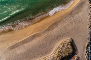 Aerial view of the Charlestown Breachway in Rhode Island with green water, surf, and tire tracks curving across the sand, by Mike Dooley