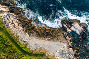 Aerial view of the Narragansett, Rhode Island coastline showing blue water, white surf, pale rock, and green grass around a rocky cove, by Mike Dooley