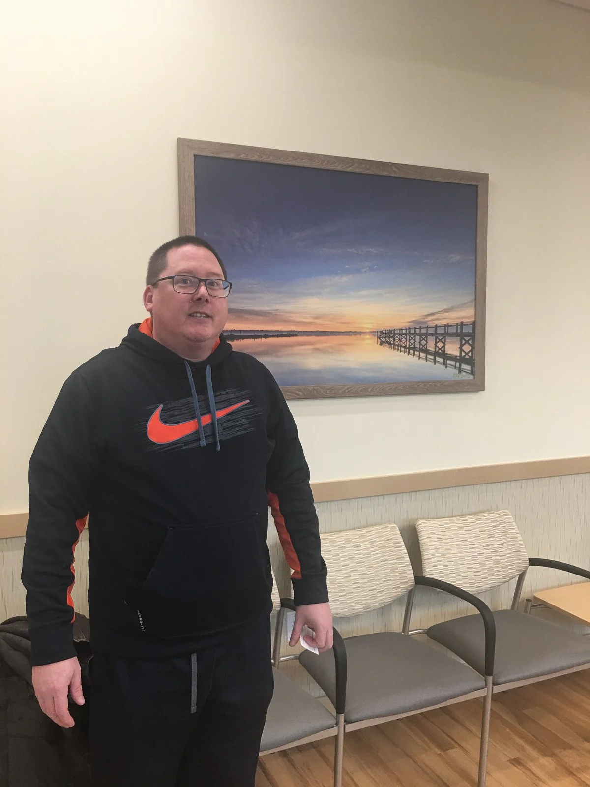 Mike Dooley standing beside his framed seascape print in a doctors' waiting room in East Providence, Rhode Island.