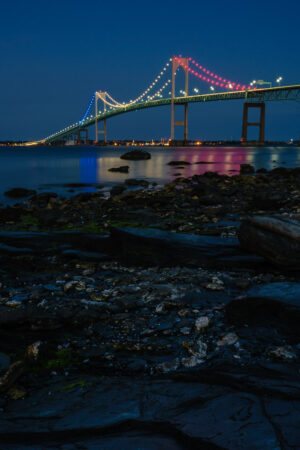 The Newport Bridge lit in red, white and blue at night above a rocky shoreline, Rhode Island, by Mike Dooley