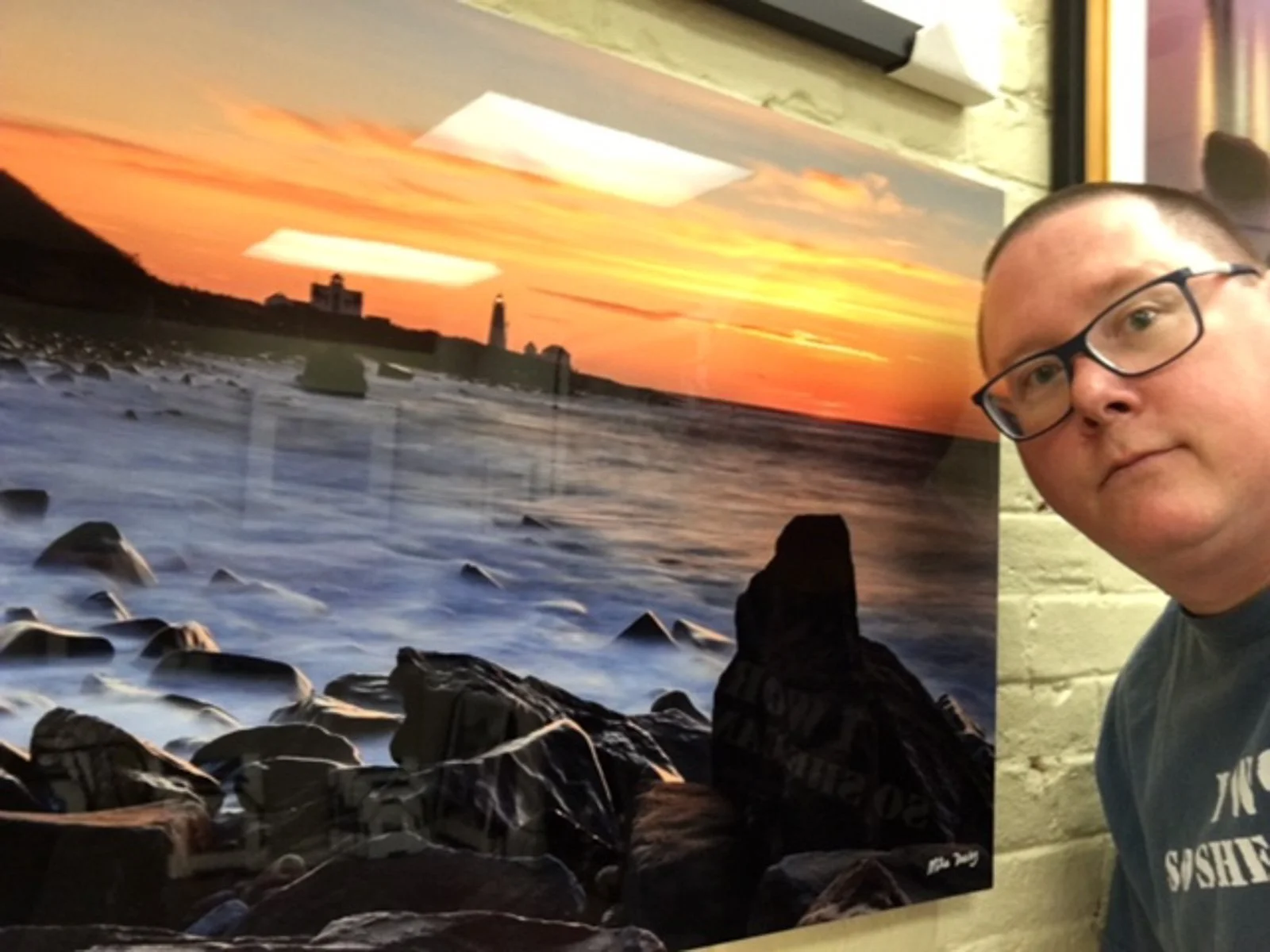 Mike Dooley next to a large metal print of Point Judith Lighthouse at sunrise.