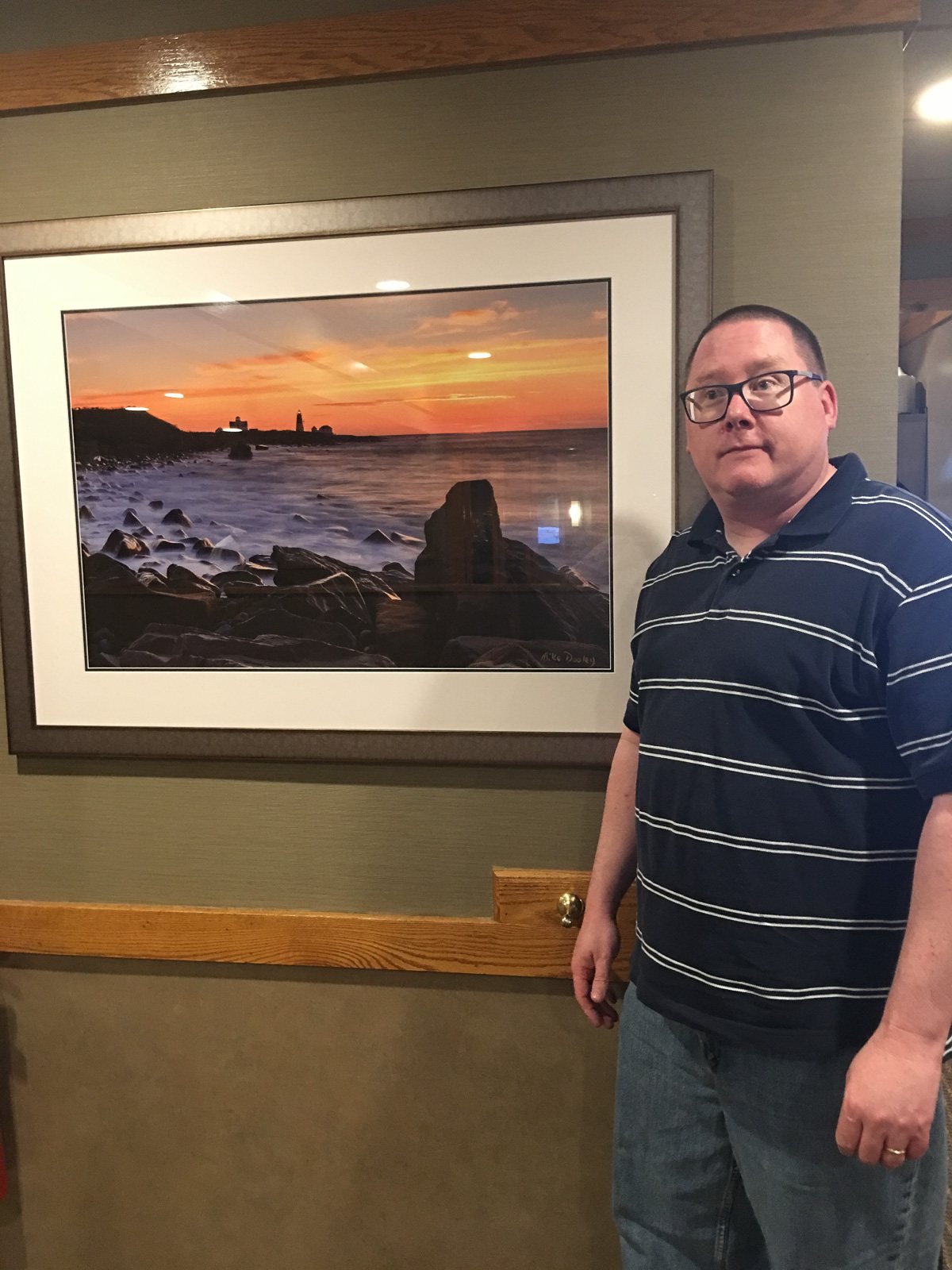 Mike Dooley standing beside his large framed print of Point Judith Lighthouse at dawn hanging in a local restaurant.
