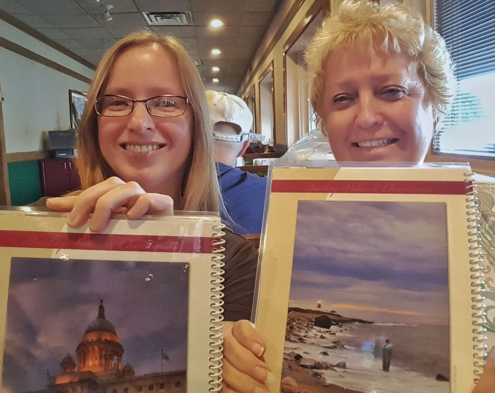 Two women in a restaurant holding up menus that feature Mike Dooley's photographs of the Rhode Island State House and a lighthouse.