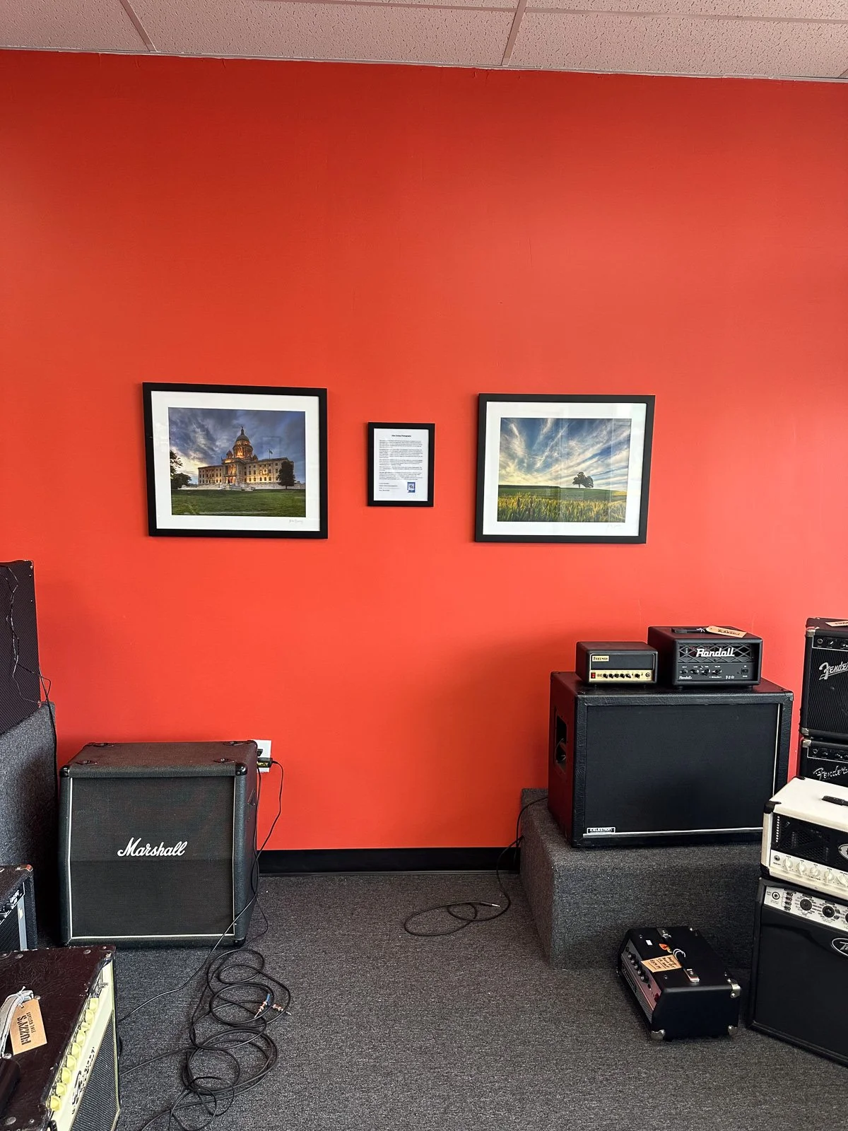 Two framed Mike Dooley prints, the Rhode Island State House and Two Trees on a Hill, hanging on an orange wall in a local music shop.