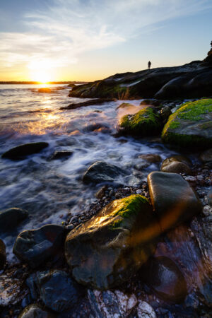 A person standing on the rocks watching the sun set over the water, with surf running between mossy rocks, Beavertail, Jamestown, Rhode Island, by Mike Dooley